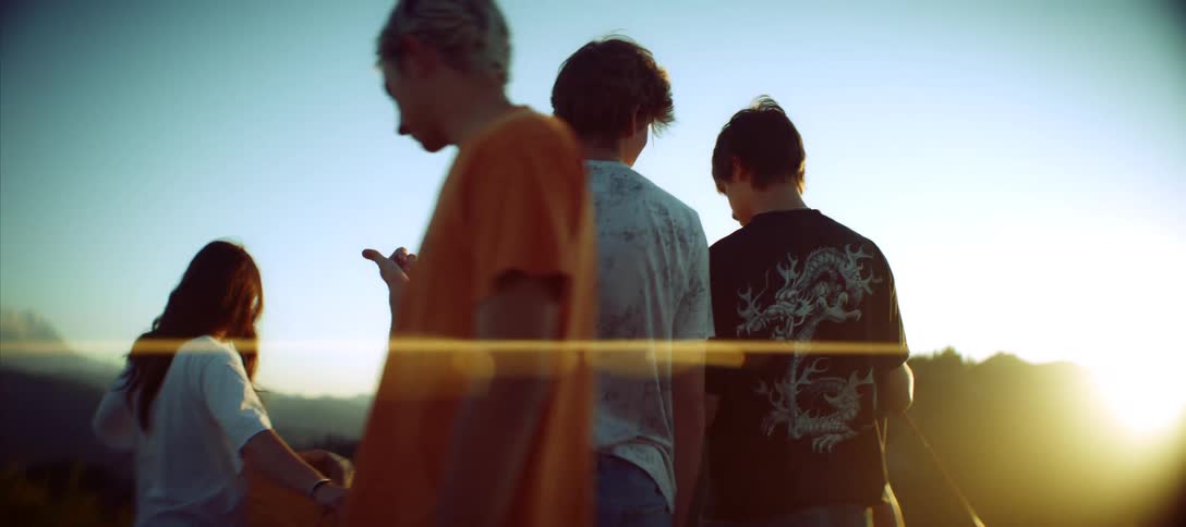 A group of young friends is standing on a scenic overlook silhouetted against a bright hazy sky the suns glow creates a lens flare as one person gestures while looking out at the distant mountains