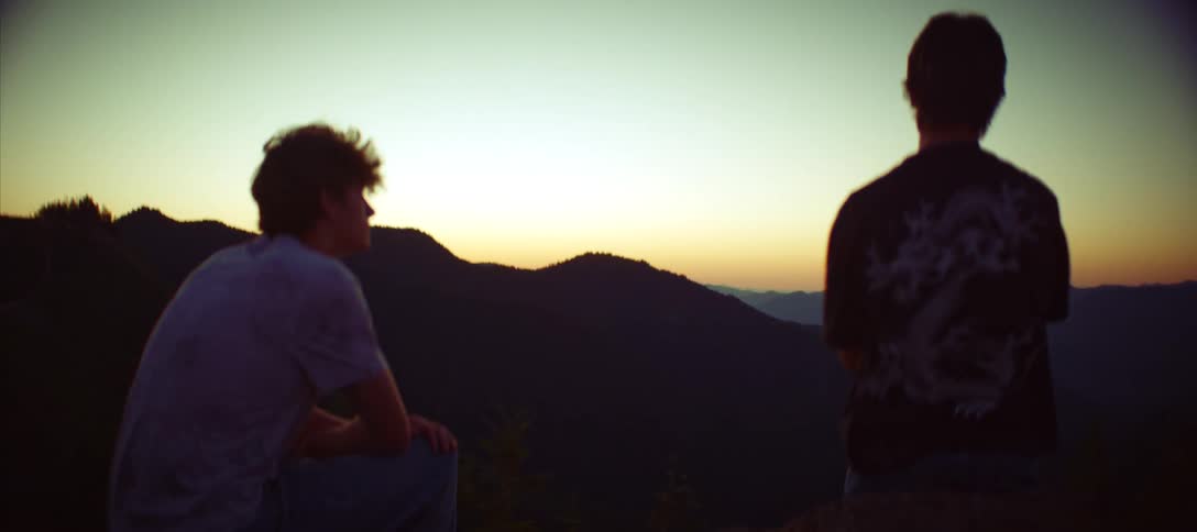 Two people stand on a mountaintop and look at the sun setting behind a mountain range