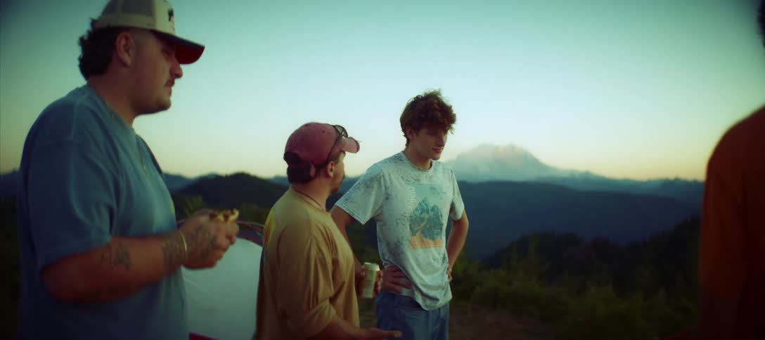 Three men are shown from the chest up talking to one another they are gathered on a hillside at dusk and a beautiful mountain is visible in the background