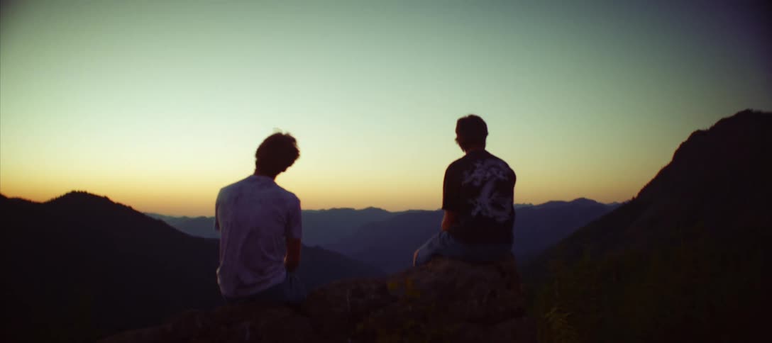 Two friends sit on a mountain ledge watching the sunset