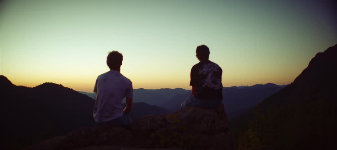 A rear view of two men sitting on a rock watching a beautiful sunset over a vast mountain range with a clear sky in a peaceful atmosphere