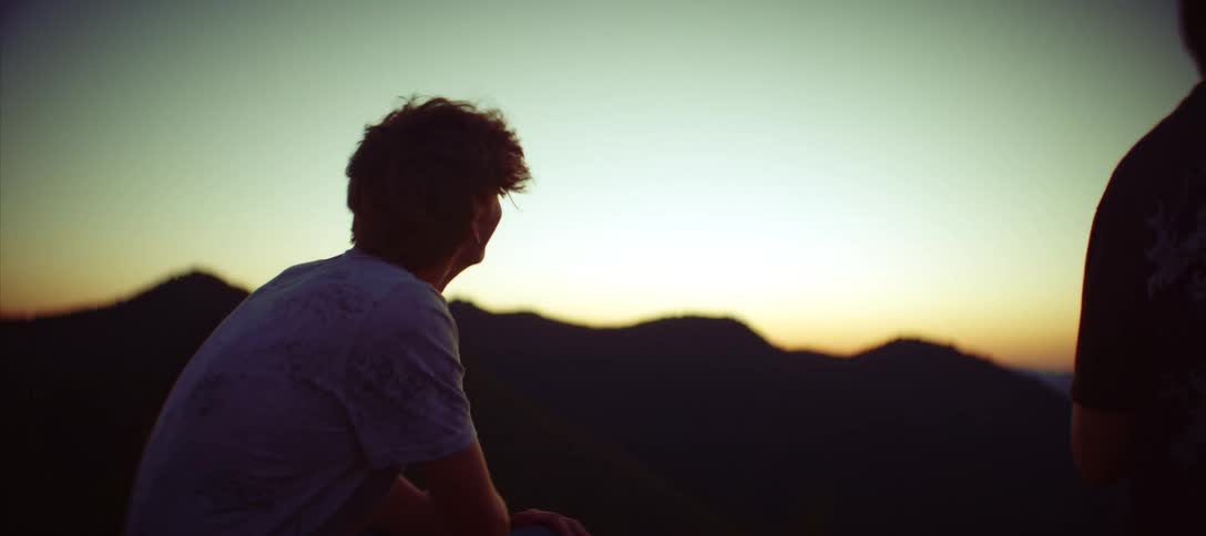 A man sits and looks out at mountains during sunset