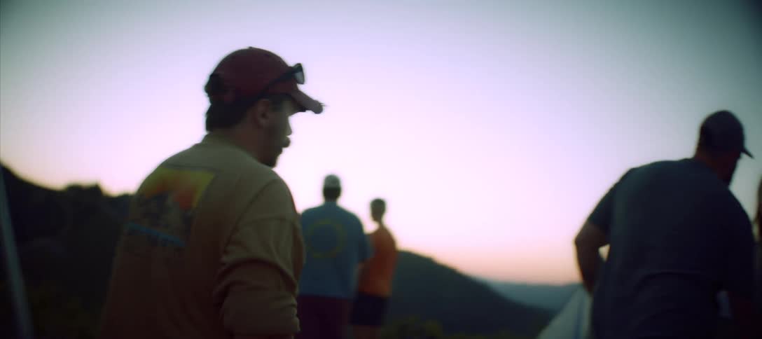 Man sitting at a campsite gets up to join a conversation with friends after dusk