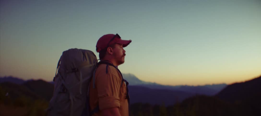 Male backpacker stands at the edge of a camp and looks out at mountainous terrain and a red horizon