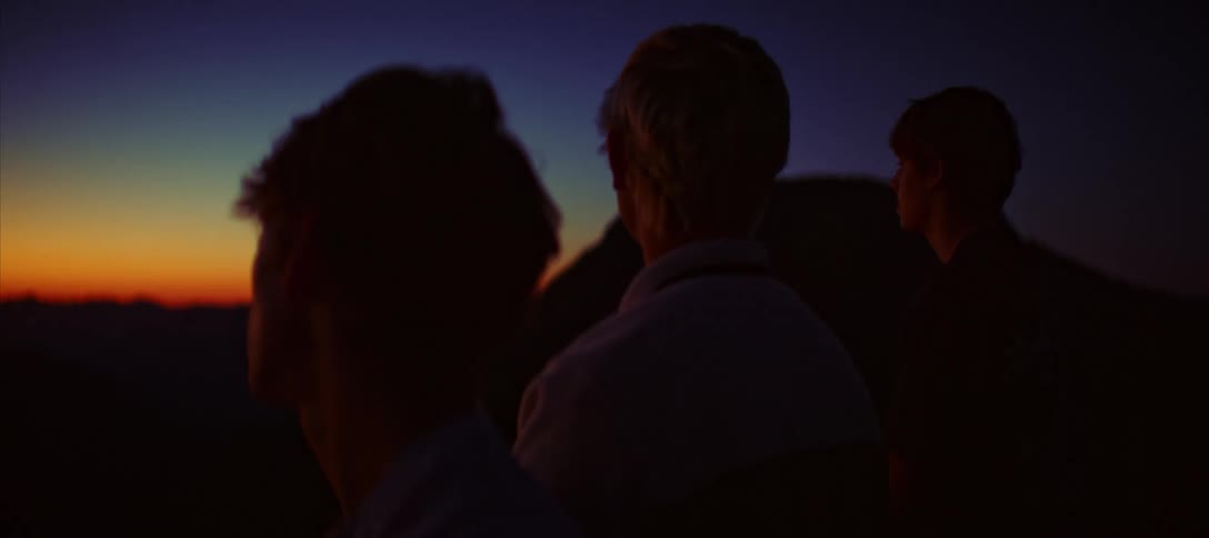 A wide shot of three people from behind with the two in the foreground in silhouette against a golden sunset in the distance a mountain range is visible against the light blue and orange sky