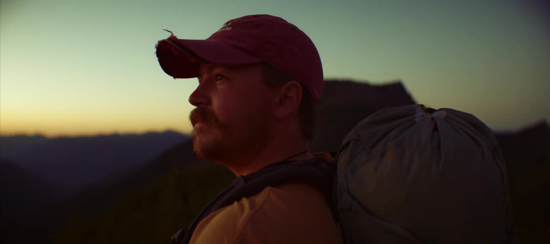 A man with backpack looks out at the mountains during sunset