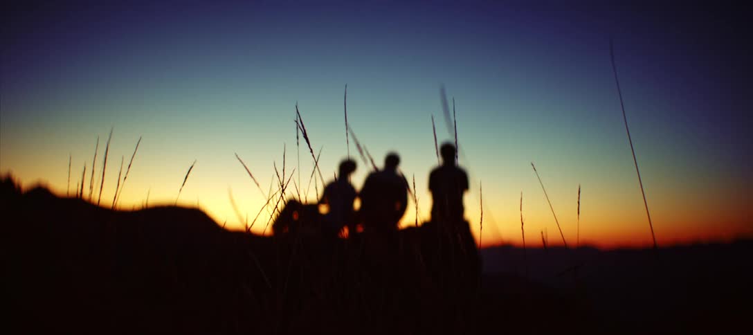 In a peaceful moment the dark silhouettes of three friends sit together on a mountain peak against a vibrant orange and blue sunrise before the focus slowly shifts to the blades of wild grass