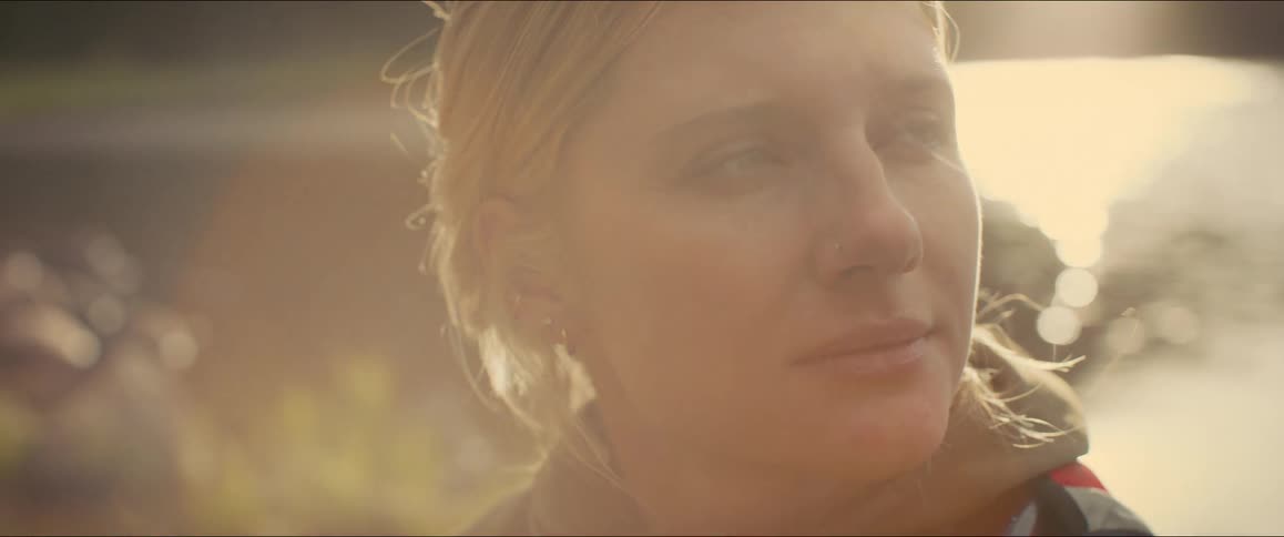 A closeup shot shows a blonde woman with her hair tied back looking offscreen with a somber expression the background is a sundrenched blurry expanse of what appears to be a lake