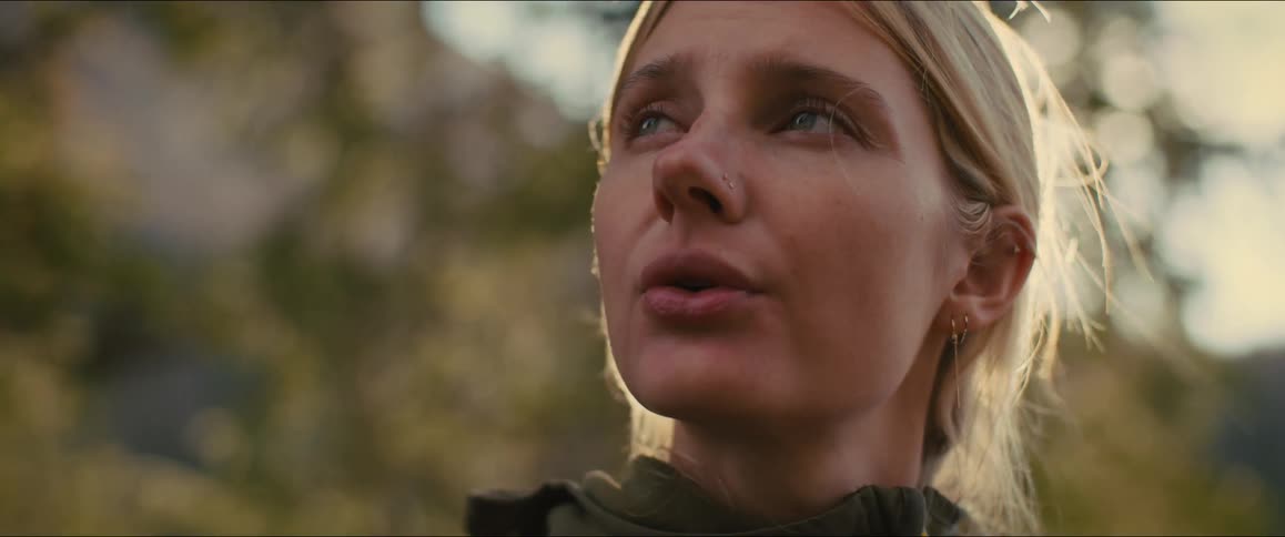 Blonde woman standing in a forest near the mountains on a sunny day looks around and takes a deep breath