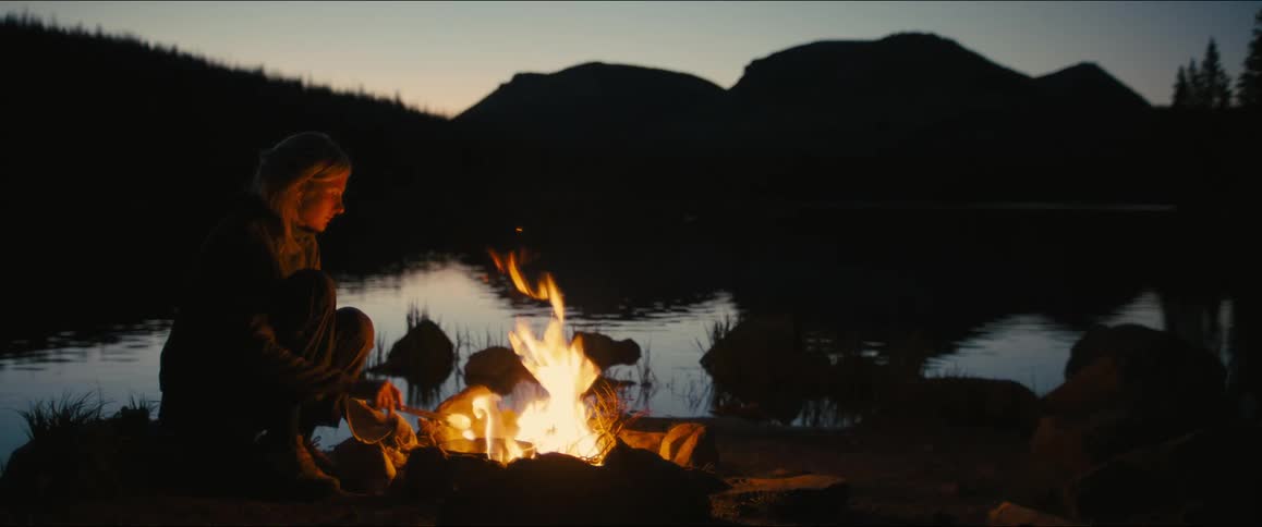 A person cooks over a campfire by a mountain lake at dusk