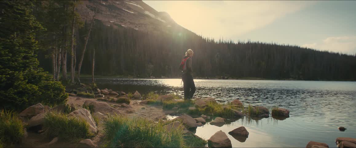 Female hiker crouches on the shore and looks across the water towards the forest at the base of a mountain on a cloudy autumn day