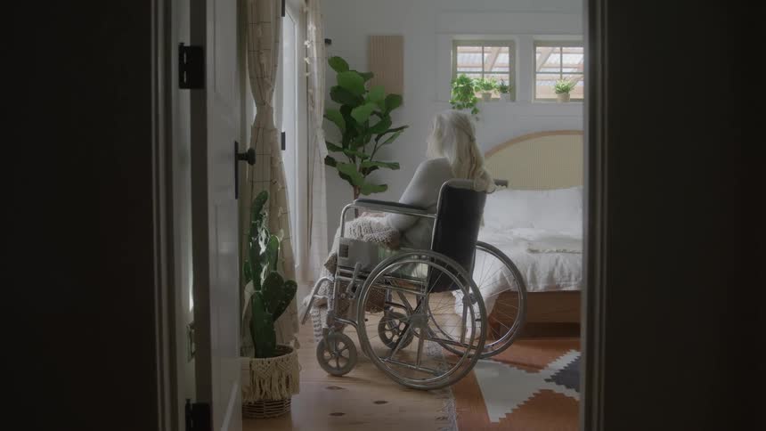 A view from a dark hallway shows an elderly person with long white hair sitting in a wheelchair they are facing a bed in a bright bedroom looking out a large window