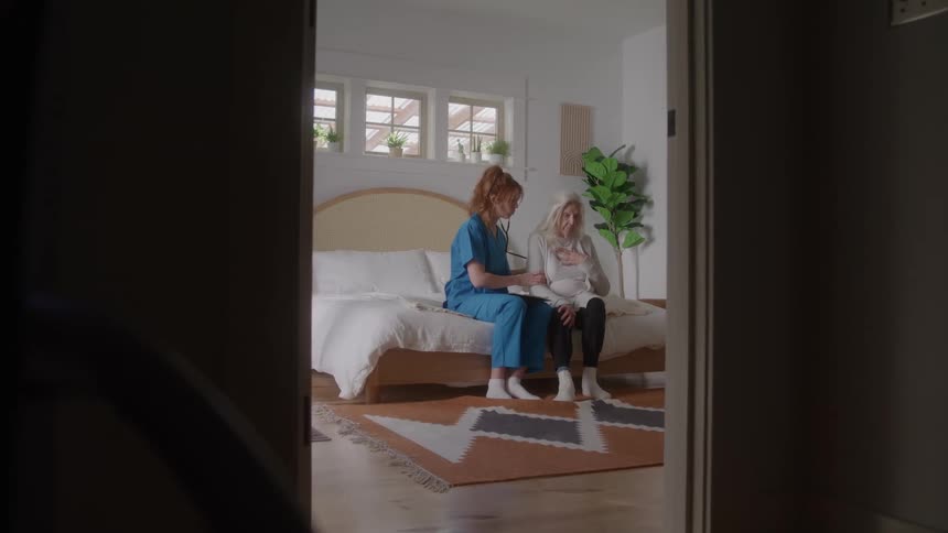 Nurse sitting on a bed beside an elderly woman in a bedroom gently encouraging her to take a deep breath