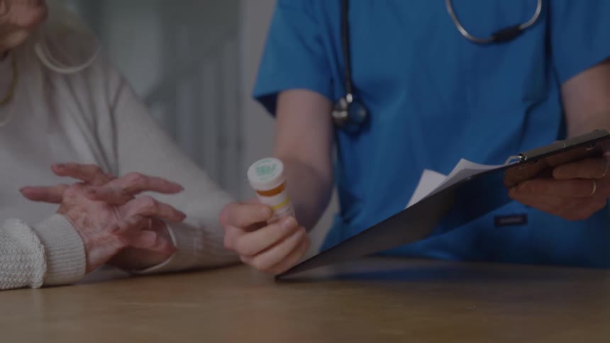 A healthcare professional sits at a table with a patient holding a clipboard and showing a pill bottle to an elderly woman