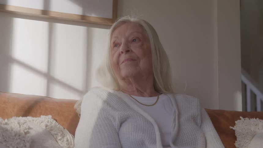 An elderly woman sits thoughtfully on a couch in a living room