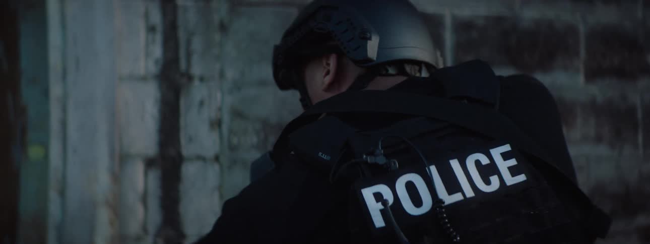 Two tactical police officers dressed in black and wearing helmets cautiously approach a building they move slowly along a grimy brick wall their firearms at the ready