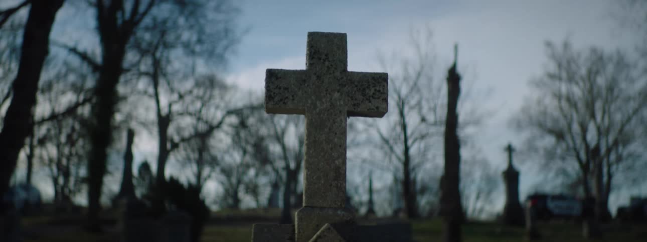 A weathered stone cross stands solemnly among graves in a quiet cemetery on a cloudy day