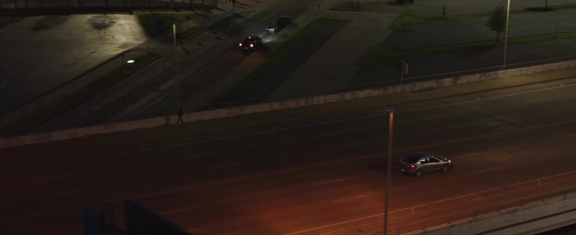 In the video an elevated shot shows a car driving on a wide empty street at dusk the camera slowly pans to follow the car as it drives away and eventually out of frame