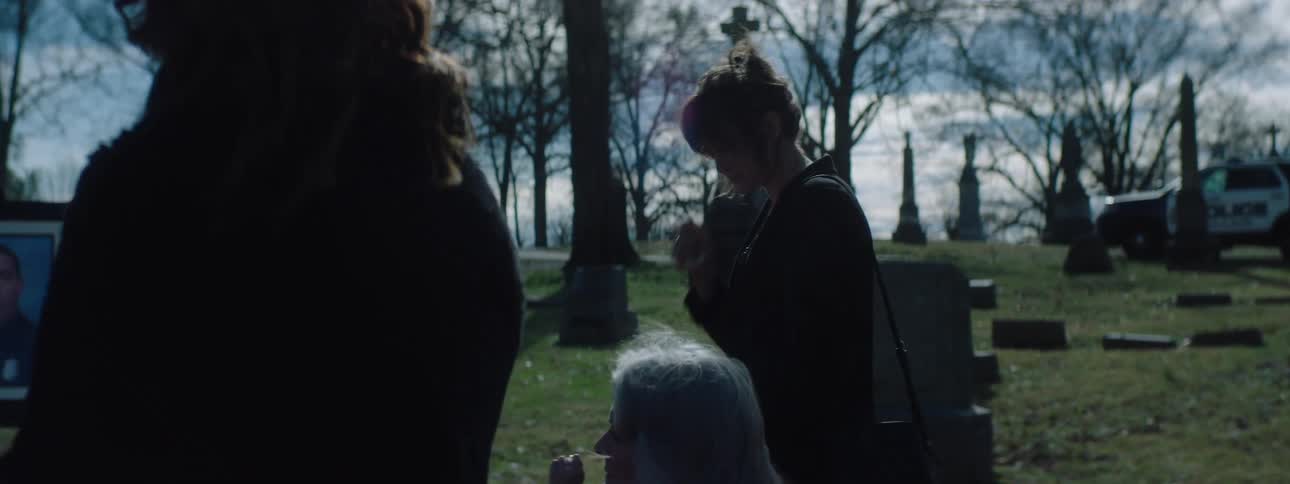 Funeral taking place in a cemetery at midday with a priest reading from his bible while standing in front of a coffin the family of the deceased stands nearby and a woman among them is shedding tears