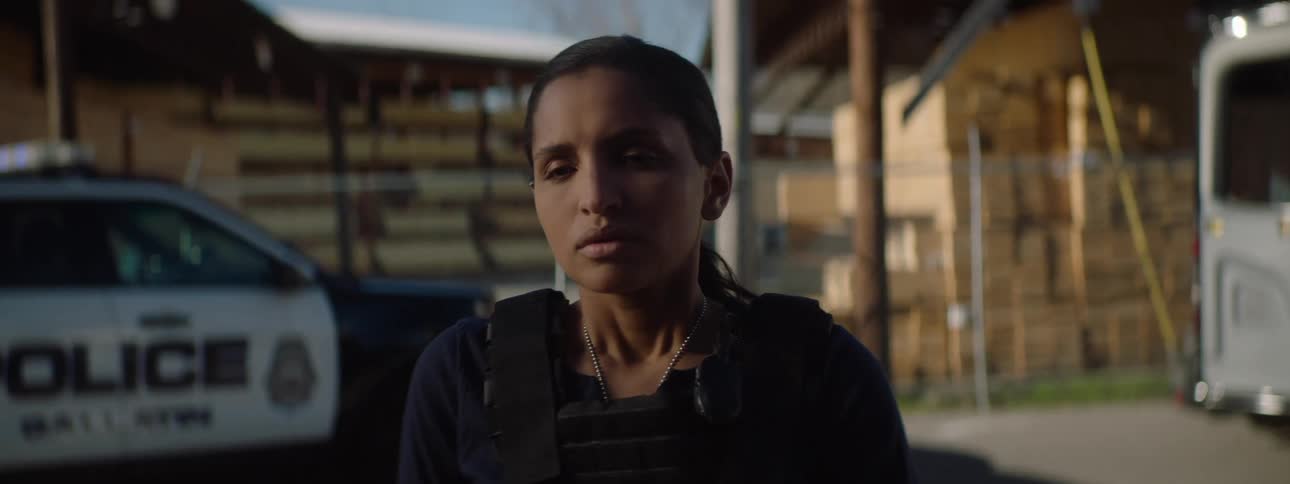 A video game scene of a seriouslooking woman in tactical gear standing near police vehicles outside a fenced industrial area