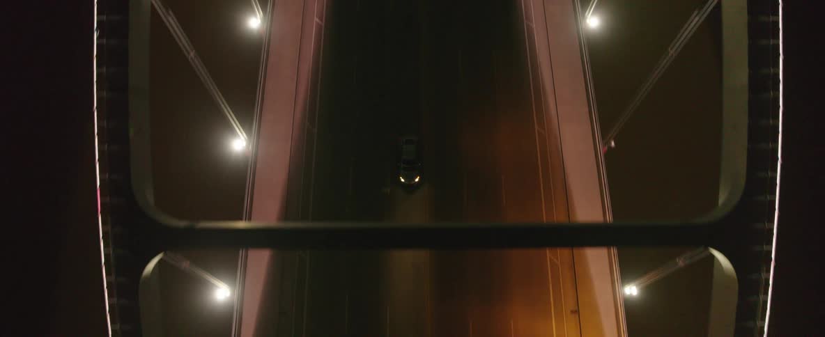 A camera rising above a bridge at night as vehicles pass underneath and the view tilts down to follow the path of the road