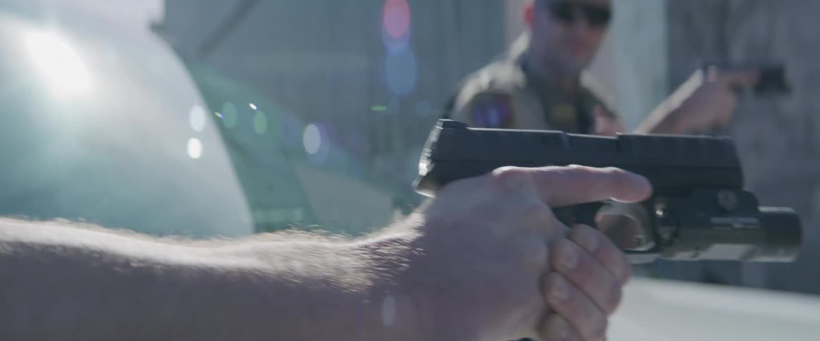 The view shifts from the determined face of a police officer to his steady hand aiming a pistol revealing another officer in the background also with his gun drawn during a tense and dangerous standoff
