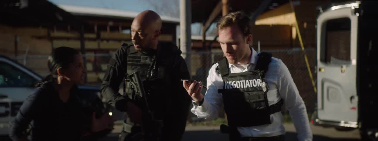 Police officers listening to the response to a negotiator express alarm while standing outdoors near police vehicles