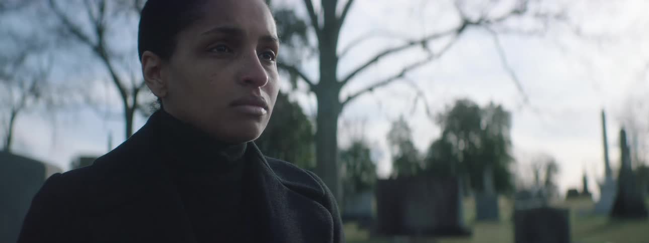 A young woman standing in a cemetery with a grief expression and some tumbstones and trees in the background