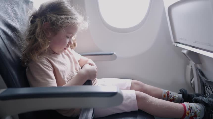 A child sits in a seat next to a window fastens a seatbelt and looks out a window