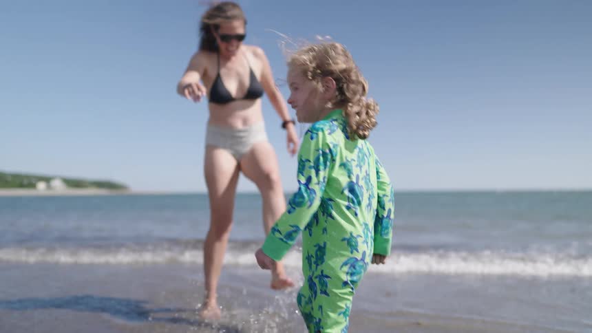 In the video a woman and a young girl are on a sandy beach near the waters edge the girl wearing a green turtlepatterned bodysuit splashes and kicks in the shallow water with a big smile on her face