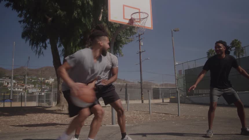 A man with a beard and dreadlocks is dribbling a basketball on an outdoor court and jumps to shoot while another man attempts to block him