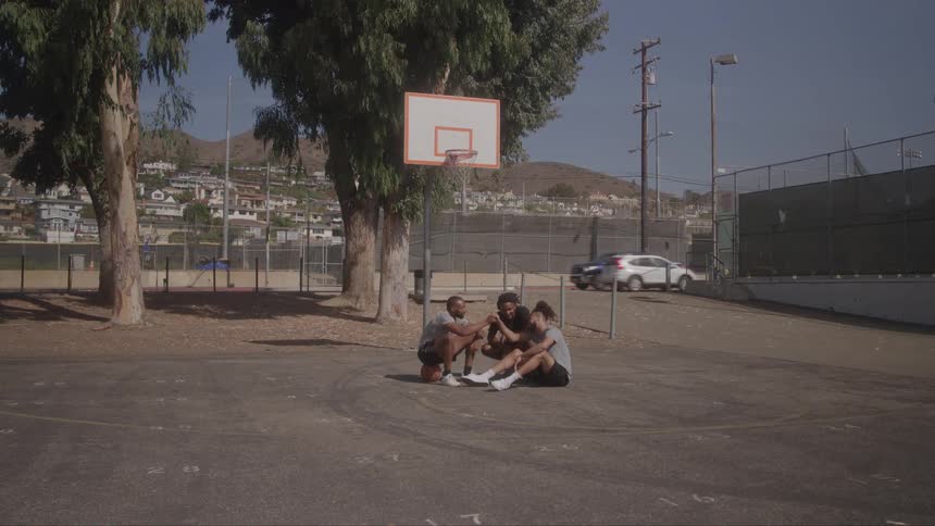 Three young men sitting on an outdoor basketball court talking under the hoop