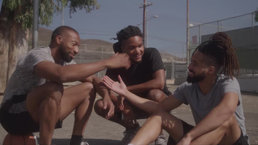 In the video three men are sitting on a basketball court with two of them squatting and one man sitting on the ground they are talking and laughing together