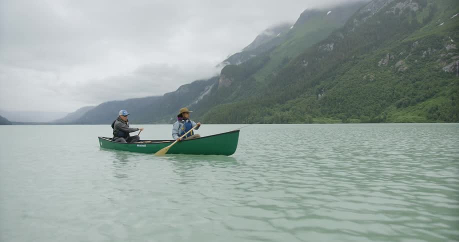 Two people are paddling a canoe across a body of water with large mountains in the background