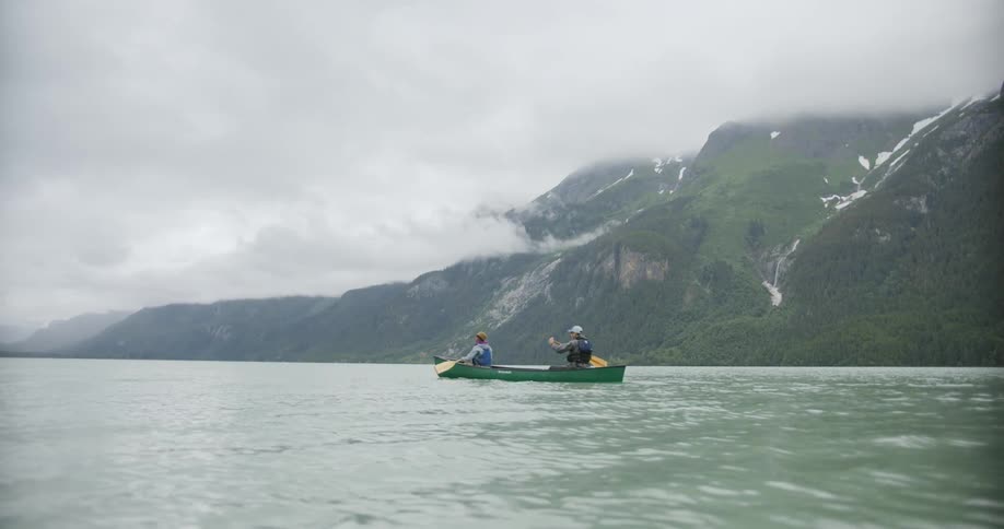 Two people are paddling a canoe across a body of water with large mountains in the background