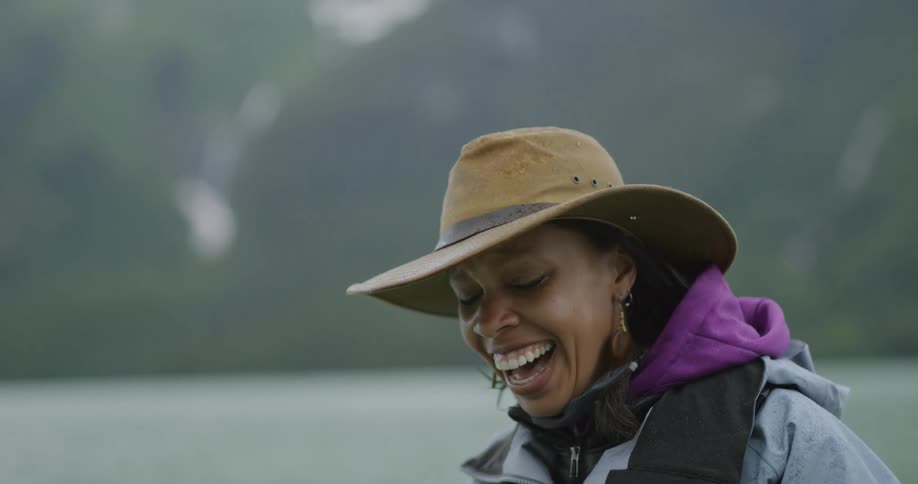 A close view shows a woman wearing a tan hat and a gray jacket with a purple hoodie underneath laughing as she looks to the side with a blurred background of mountains and a lake