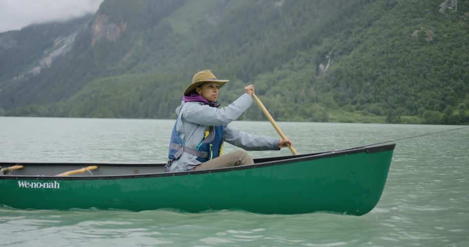 Woman in a green canoe fishing on a lake surrounded by mountains