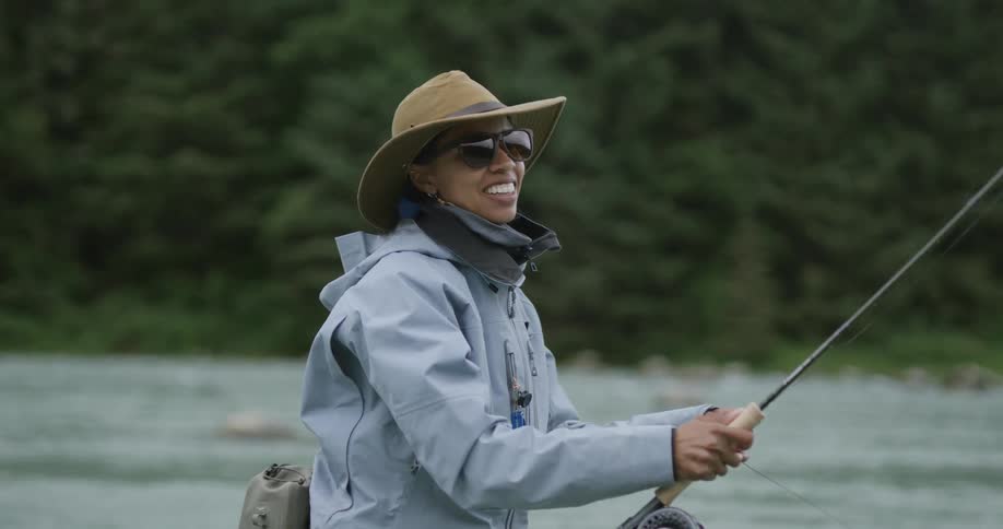 A woman wearing a tan hat sunglasses and a gray jacket is fly fishing in a river and smiling as she holds her fishing rod with a lush green forest in the background