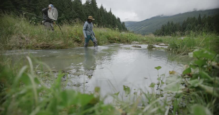 Two fishermen cross a stream surrounded by mountain vegetation
