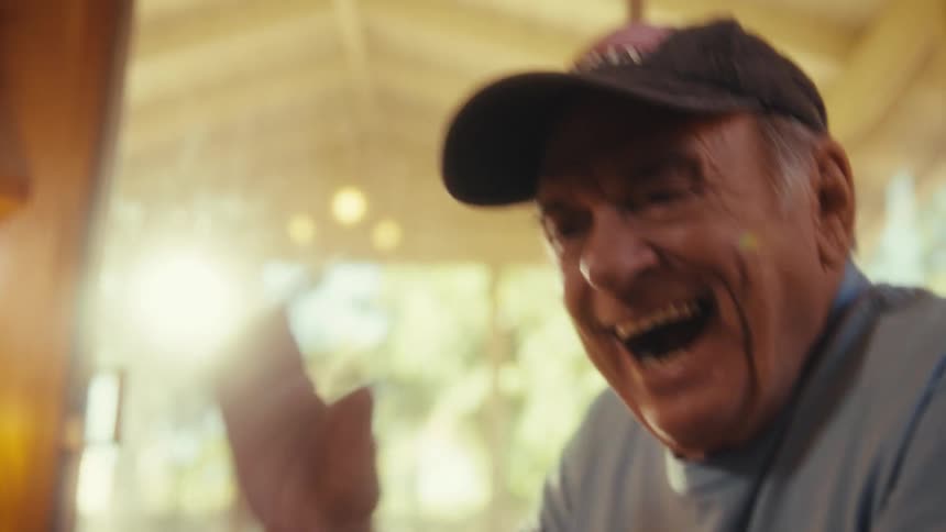 In the video a man with a veteran cap and a light blue tshirt with a star logo is sitting indoors he is holding a phone and laughing