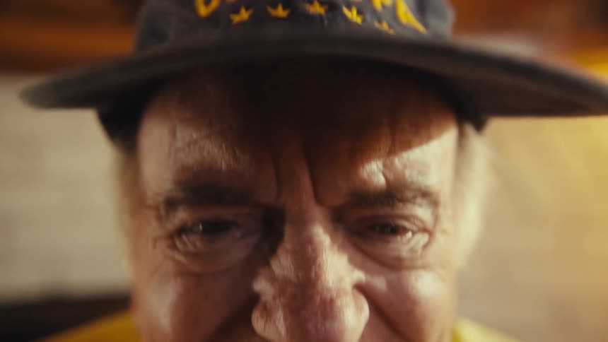 A shot from a low angle shows an elderly man wearing a black baseball cap that says worlds best grandpahe looks up at the camera and breaks into a warm smile his face lighting up