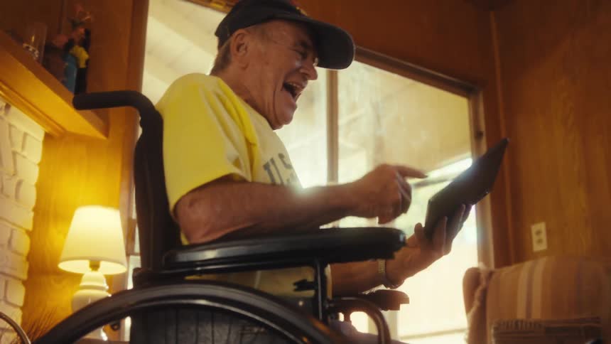Elderly man sitting in a wheelchair in his living room watching an exciting game on a tablet with full attention
