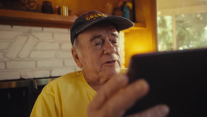 Elderly man wearing a yellow shirt and a baseball cap talking and laughing while holding a tablet on his hands indoors during day time