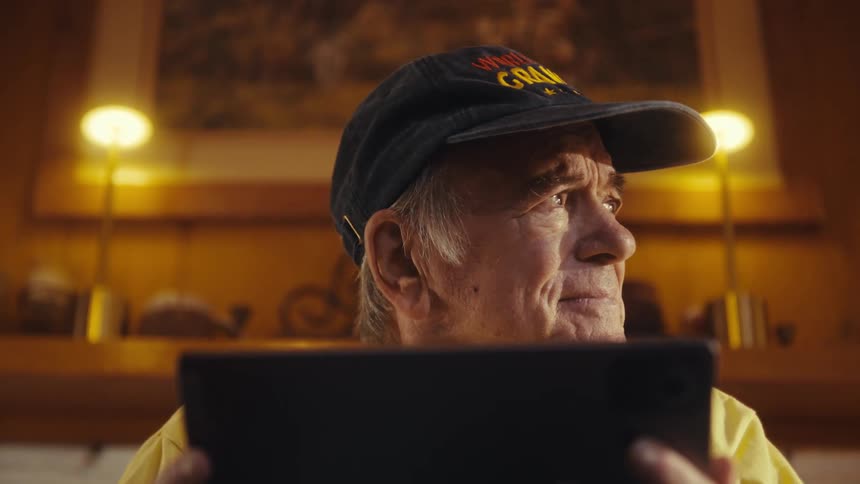 Elderly man in cap with emotional expression is sitting indoors holding a tablet