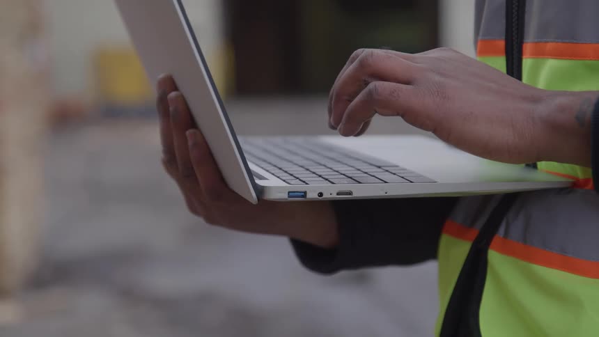 Man standing outdoors types on a laptop while holding it in one hand