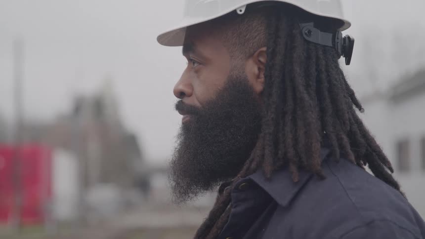Young black man with long braided hair and a full beard wearing a white hard hat standing outdoors