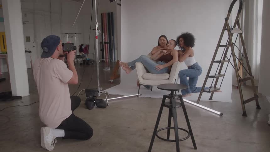 Man half kneels to take video of three women posing with a chair in a studio during the daytime