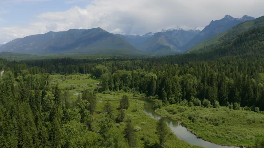 A breathtaking aerial shot slowly pulls backward to reveal an epic and vast mountain landscape featuring a lush green valley with a meandering river dense evergreen forests and a majestic snowcapped range