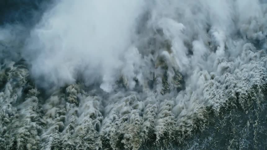 A powerful waterfall cascades down a cliff and crashes into the dark water below creating a huge plume of white spray and mist the churning water at the base of the falls is a mix of foamy white and deep grey
