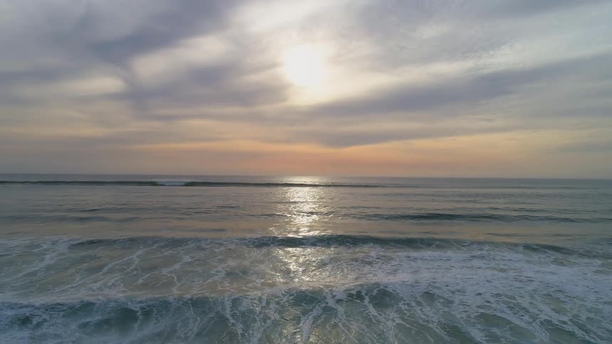 A beautiful lowaltitude aerial shot flies backward over the ocean towards the shore capturing the suns golden reflection shimmering across the surface as gentle waves with white foam roll in at sunset